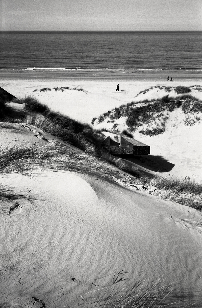 Photographie en noir et blanc d'une plage du Nord de la France, montrant des dunes de sable avec de l'herbe, une structure abandonnée et des personnes se promenant au bord de la mer.