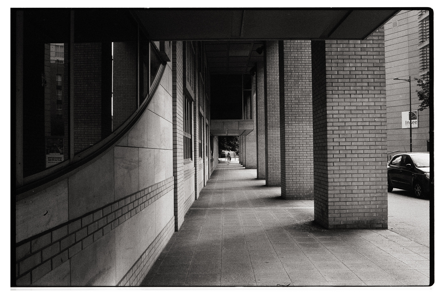 Un couloir urbain en noir et blanc avec des colonnes en brique, éclairé par une lumière douce, où un passant solitaire marche au loin.