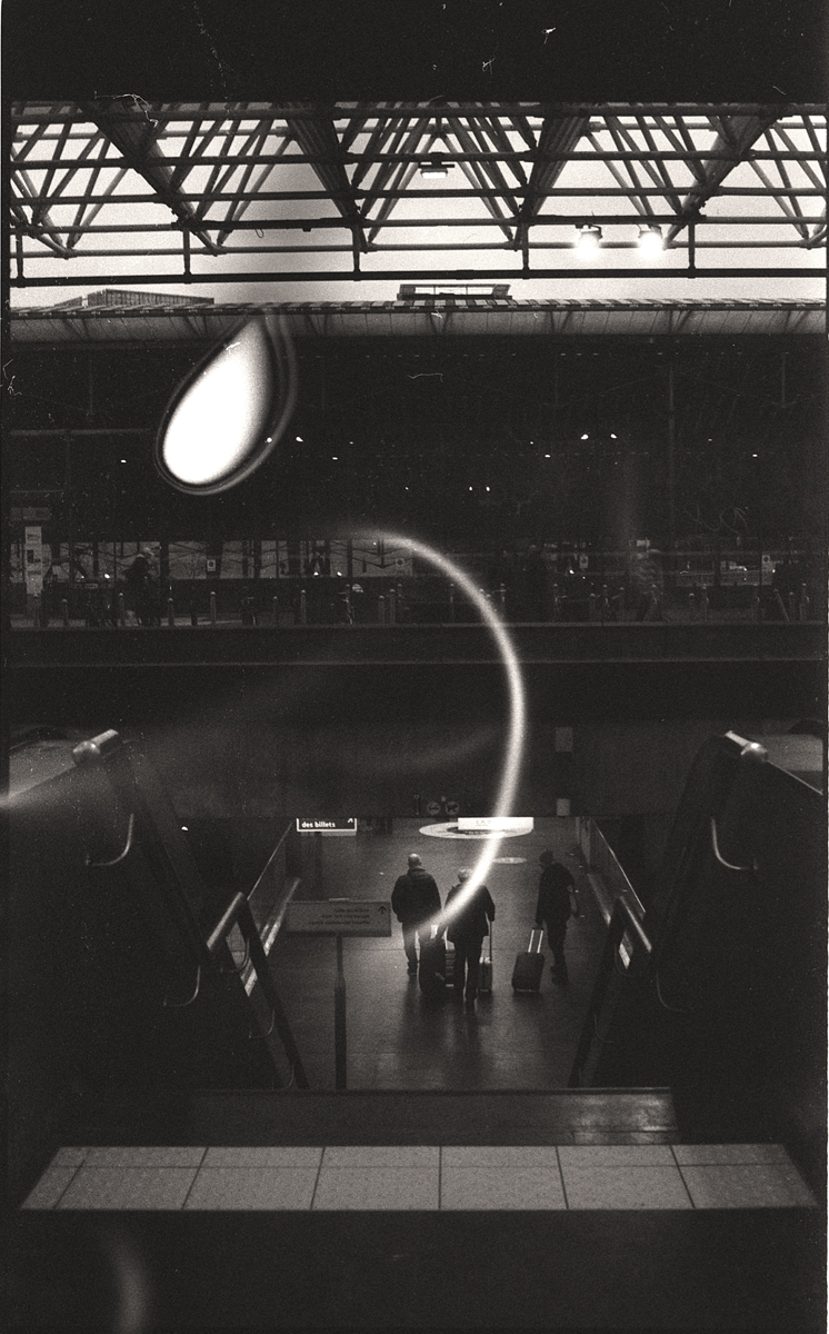 Photographie en noir et blanc d'une gare, montrant un mouvement flou de passagers descendant des escaliers, avec un éclairage dramatique et des éléments architecturaux en arrière-plan.