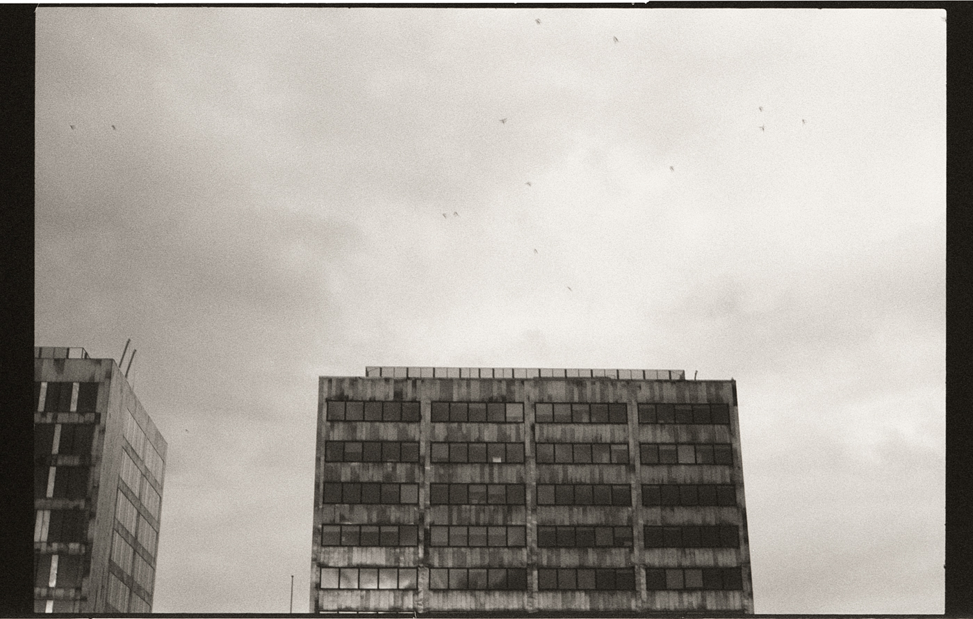 Photographie en noir et blanc de deux immeubles de bureaux, avec un ciel nuageux en arrière-plan. L'image capture des détails architecturaux et des textures, mettant en évidence la verticalité des structures.