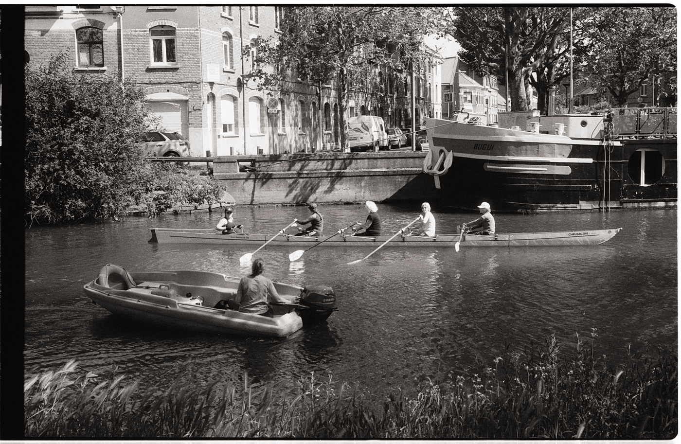 Des kayakistes s'entraînant sur un canal en été, avec un bateau en arrière-plan et des bâtiments en bordure.