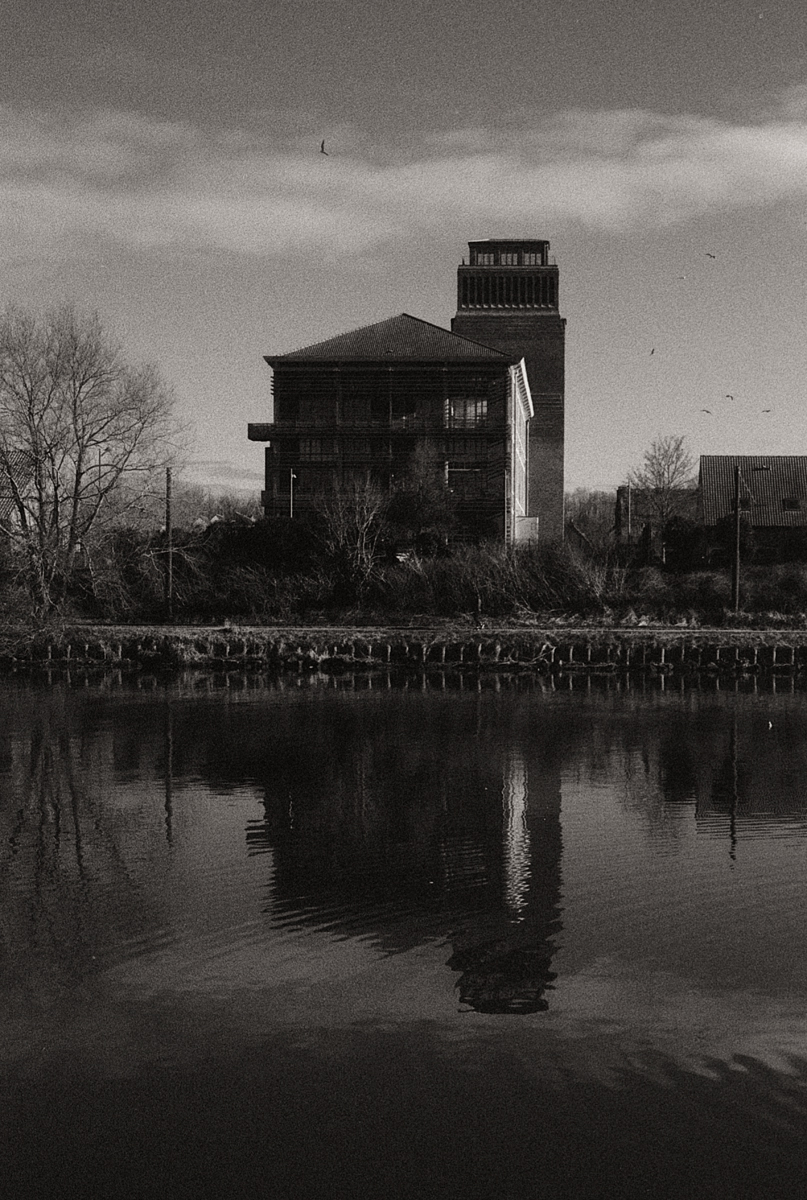 Une maison sombre se reflète dans l'eau calme d'un canal, avec des arbres au premier plan et un ciel nuageux en arrière-plan.