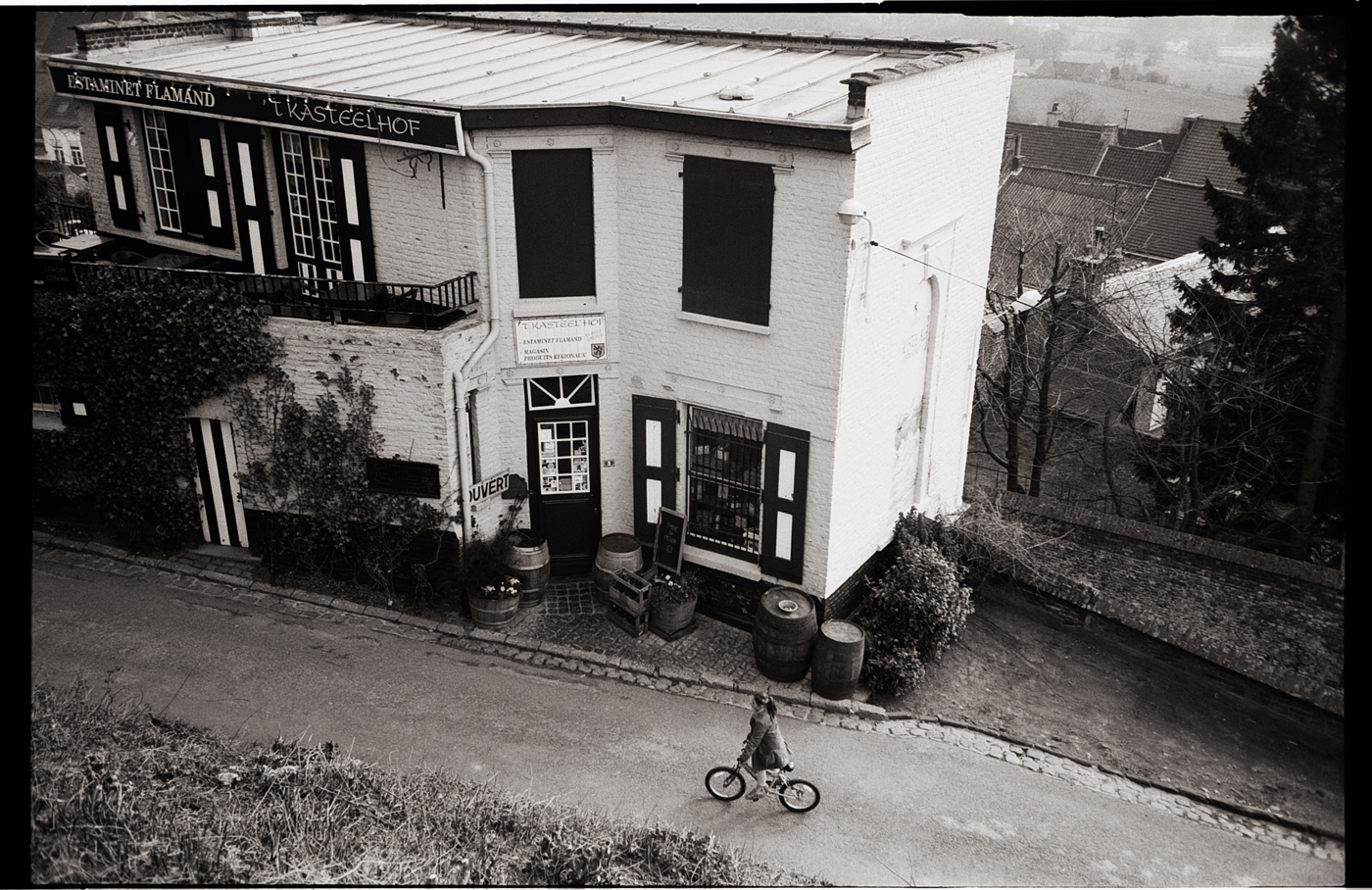 Un enfant à vélo passant devant un bâtiment en briques blanches, avec des fenêtres à volets noirs, dans une scène urbaine en noir et blanc.