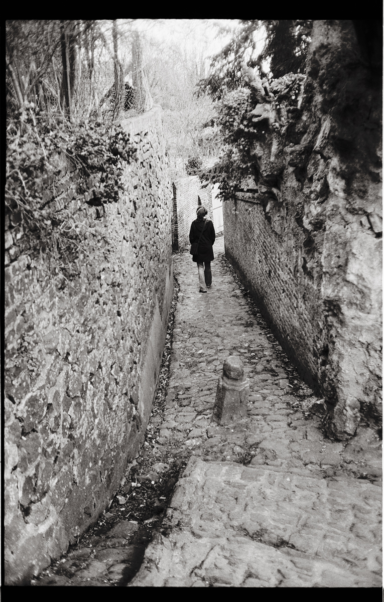 Une personne marchant seule dans un ru étroit, entourée de murs en pierre et de végétation, capturée en noir et blanc.