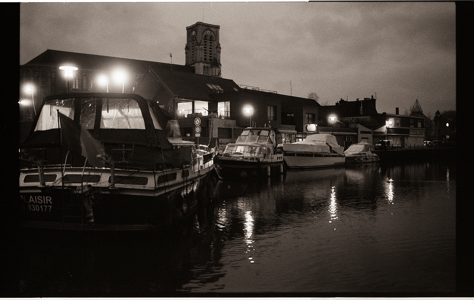 Photographie nocturne en noir et blanc montrant des bateaux amarrés le long d'un quai, avec un bâtiment éclairé en arrière-plan dans une ambiance paisible.