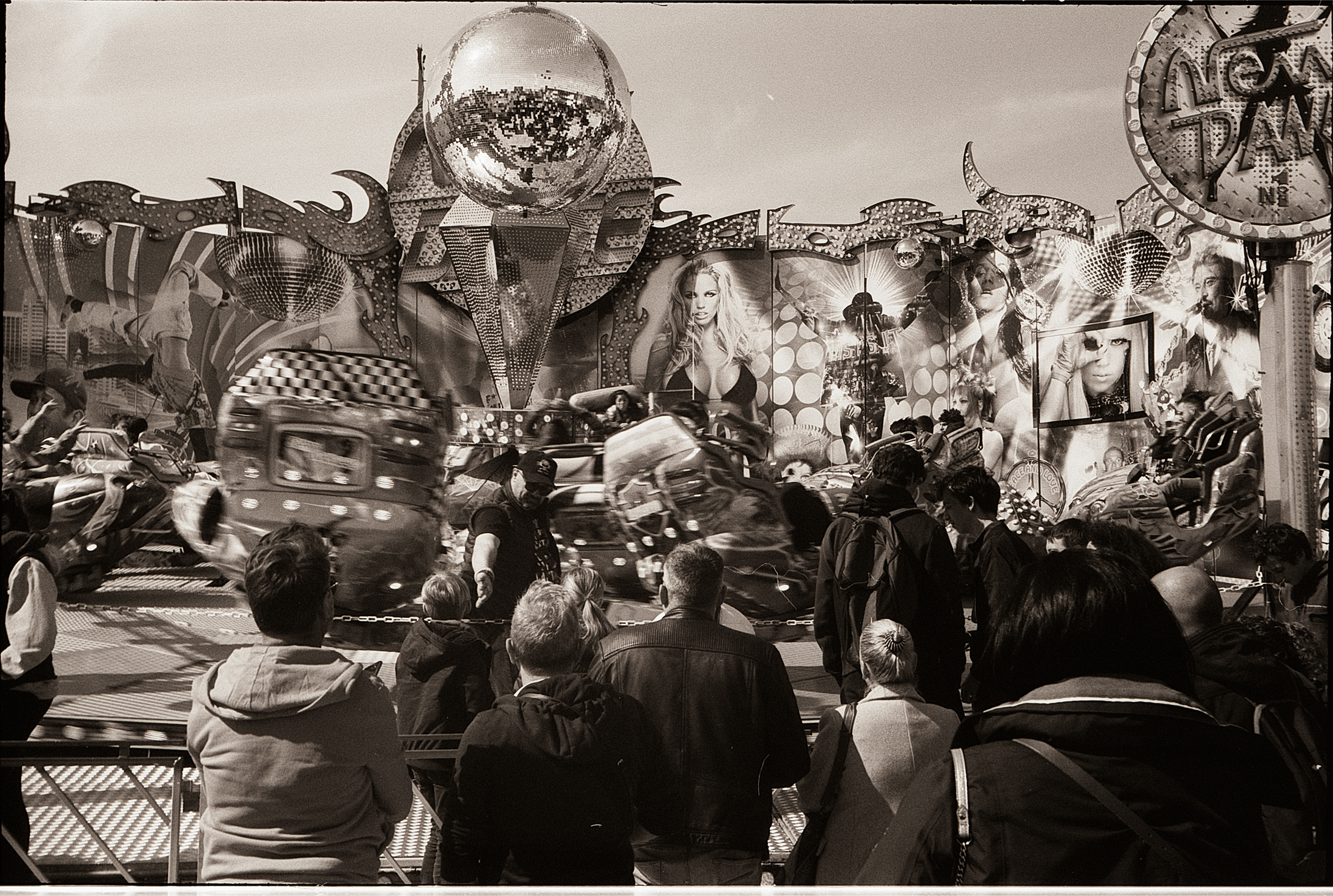Une fête foraine animée à Lille, capturée en noir et blanc, avec des manèges en mouvement et une foule rassemblée devant des décorations colorées.