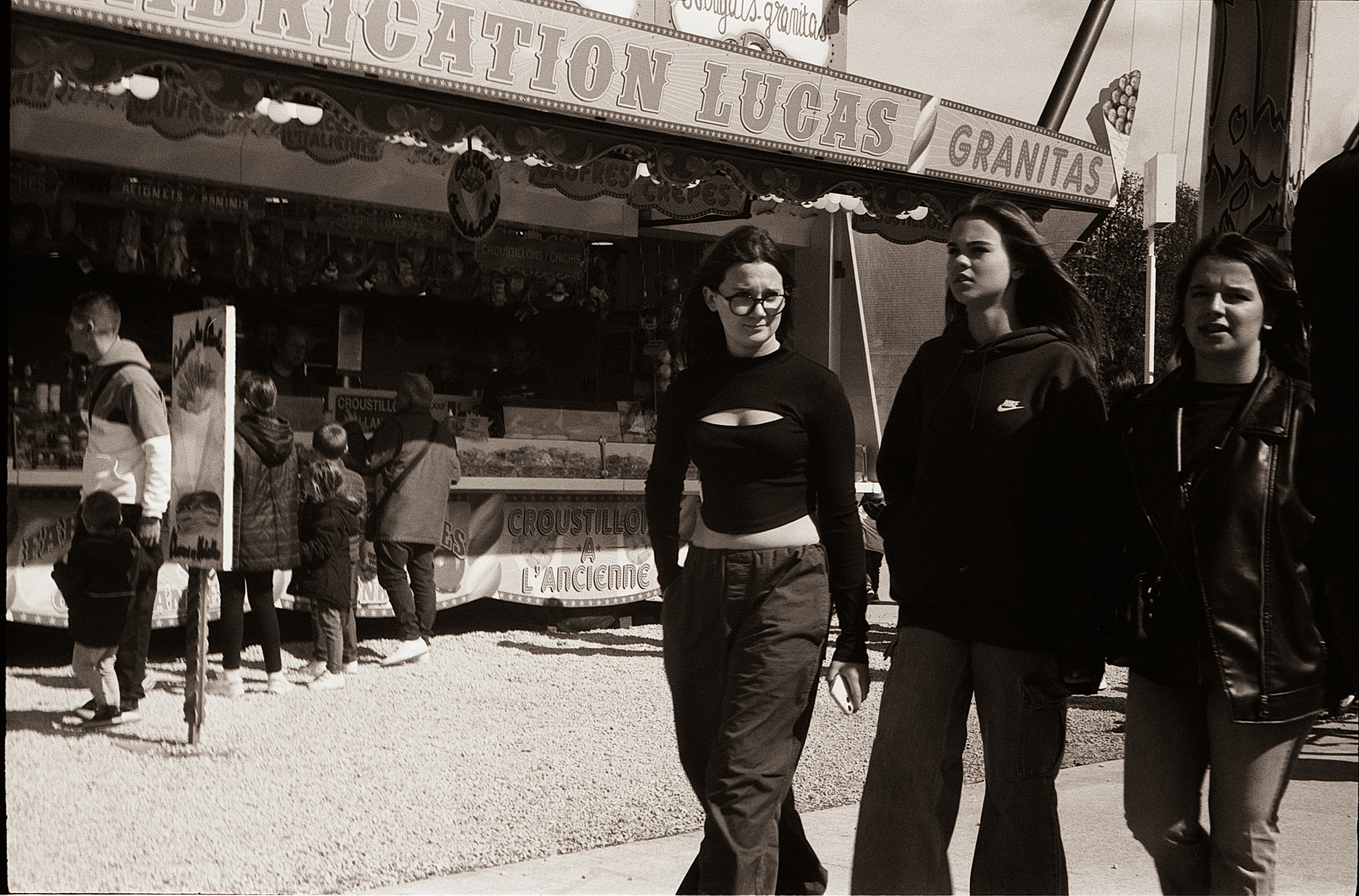 Trois jeunes femmes marchant devant un stand de nourriture au bord d'une fête foraine, avec des enfants en arrière-plan.