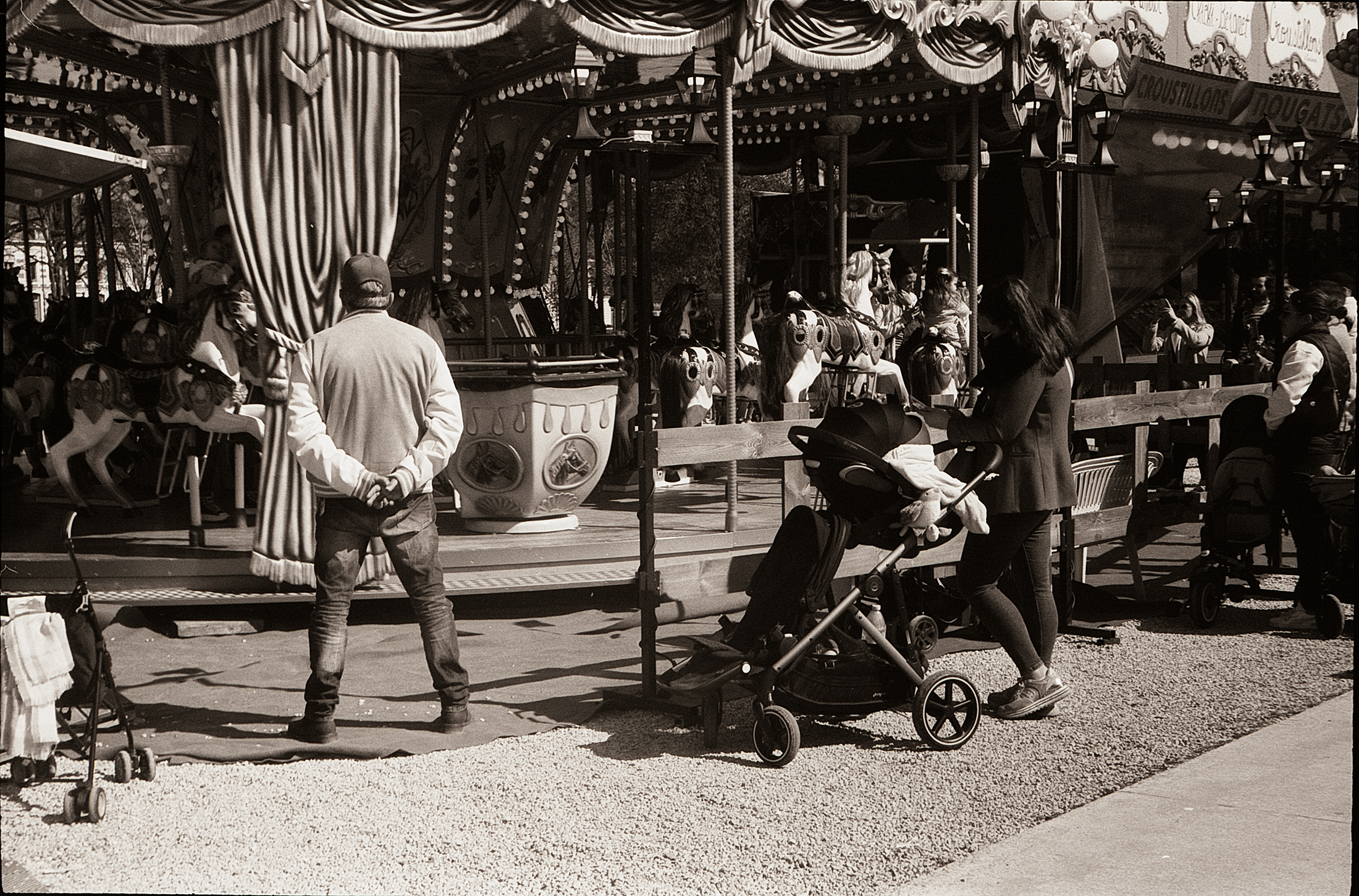 Un homme de dos observe un carrousel lors d'une fête foraine, avec des enfants et des familles autour, en ambiance argentique.