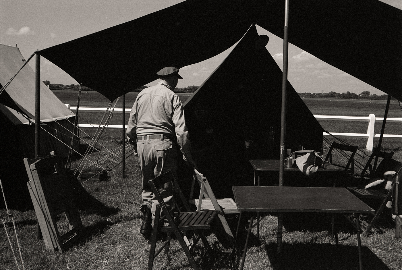 Un homme en uniforme militaire marche vers une tente sous un ciel partiellement nuageux, avec des tables et des chaises en bois à proximité.