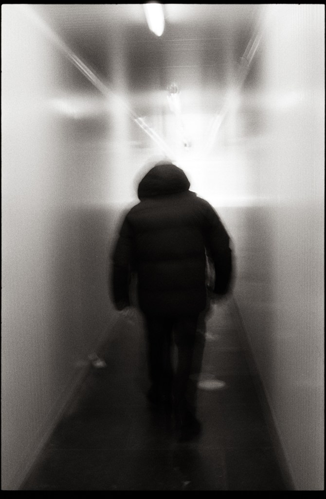 Un individu marchant à travers un couloir flou, portant un manteau noir, dans une ambiance monochrome. Photographie de rue floue.