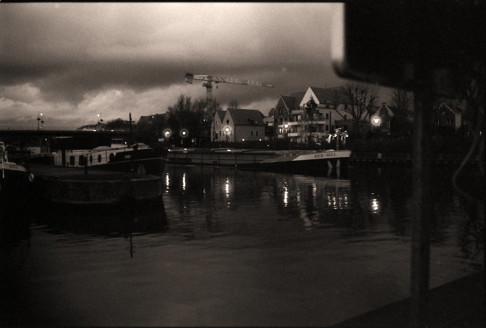 Photographie argentique noir et blanc du port de plaisance de Wambrechies au crépuscule hivernal. Une péniche traverse l'arrière-plan tandis que des lampadaires allumés créent des reflets chaleureux dans l'eau. L'image présente un grain prononcé et un flou atmosphérique caractéristiques de la pellicule, capturant une ambiance mélancolique aux tons bruns.