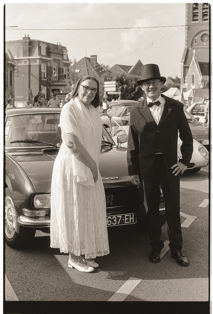 Un couple élégant se tenant devant une voiture vintage, posant pour la photographie en noir et blanc, avec un homme en costume et un haut de forme, et une femme en robe blanche.