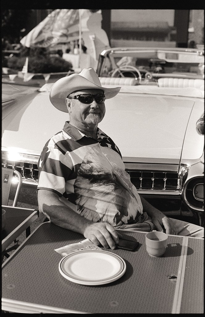 Un homme souriant portant un chapeau de cowboy et des lunettes de soleil, assis à une table avec des assiettes et une tasse en plastique, devant une voiture classique.