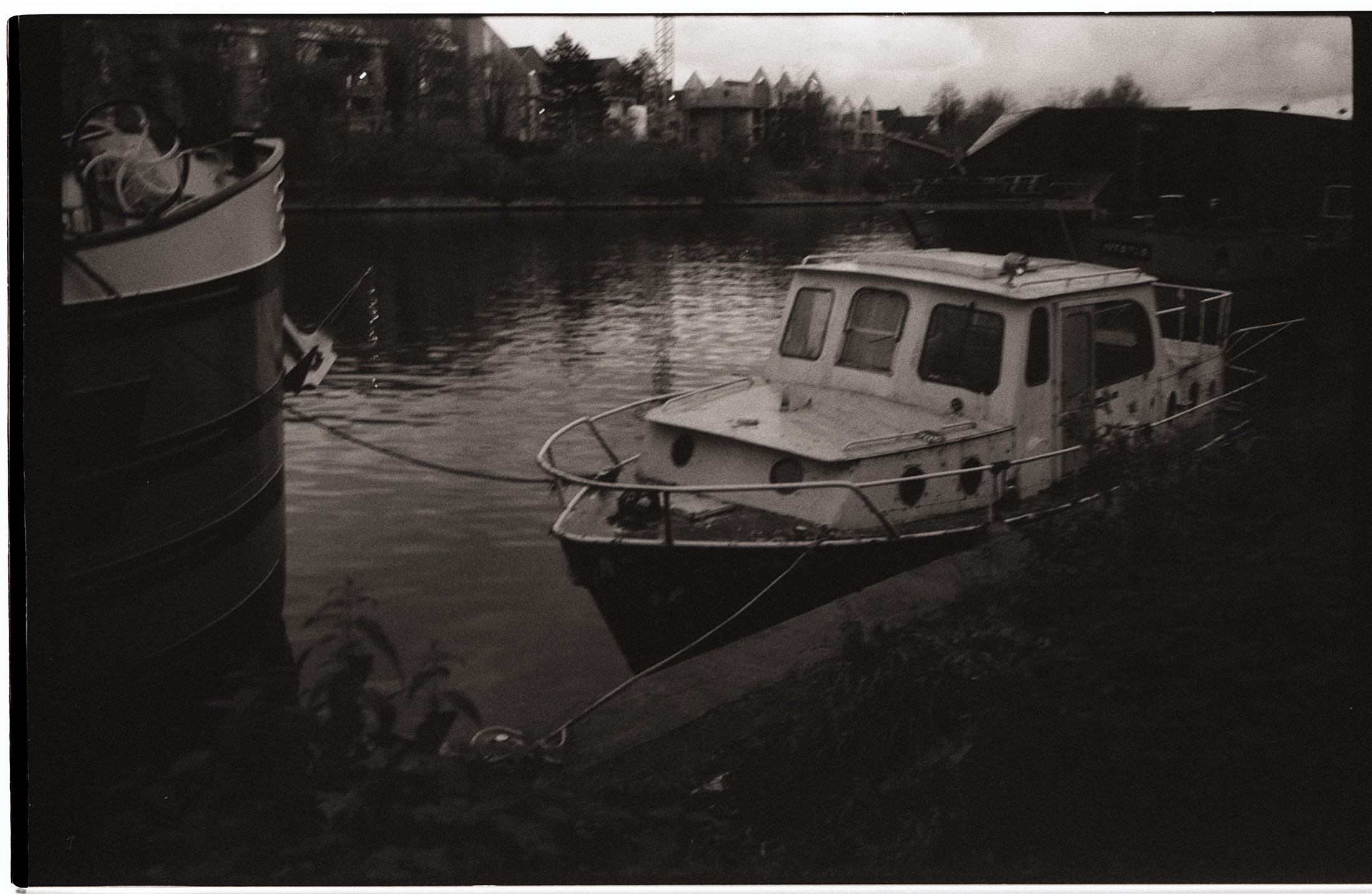 Un bateau amarré près d'une berge, photographié en noir et blanc, avec des détails flous et un grain prononcé, créant une ambiance nostalgique.