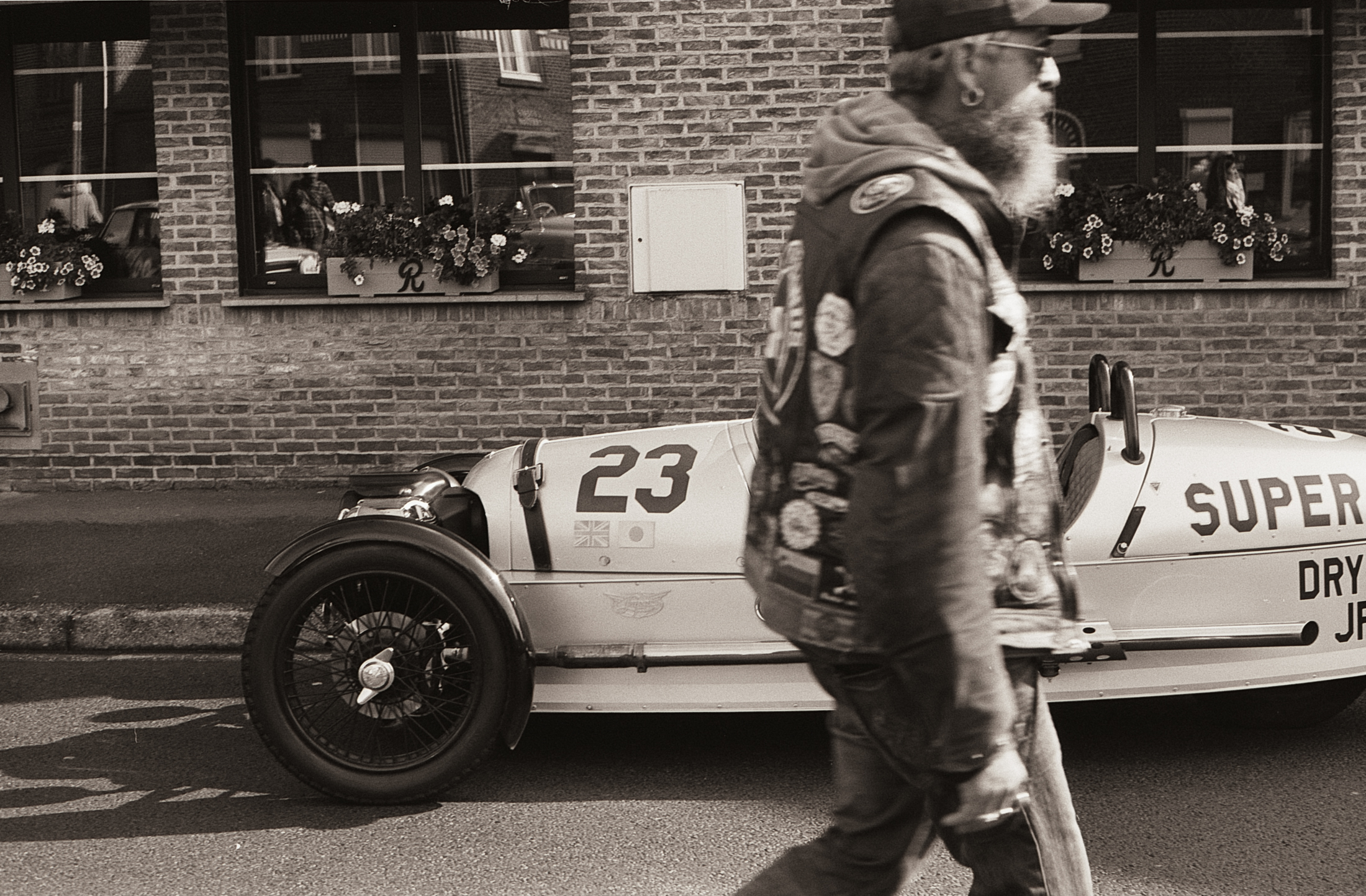 Un homme avec une barbe et un gilet décoré de patchs marche à côté d'une voiture vintage dans une rue pavée, avec des fenêtres en briques et des fleurs en arrière-plan.