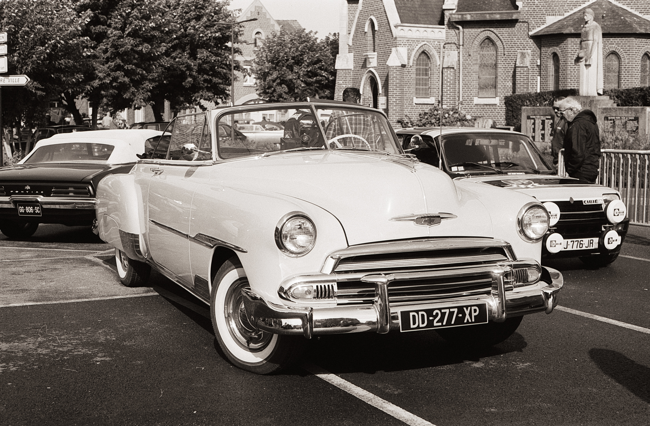 Une voiture convertible blanche des années 1950 garée dans une rue, entourée d'autres véhicules et d'arbres en arrière-plan.