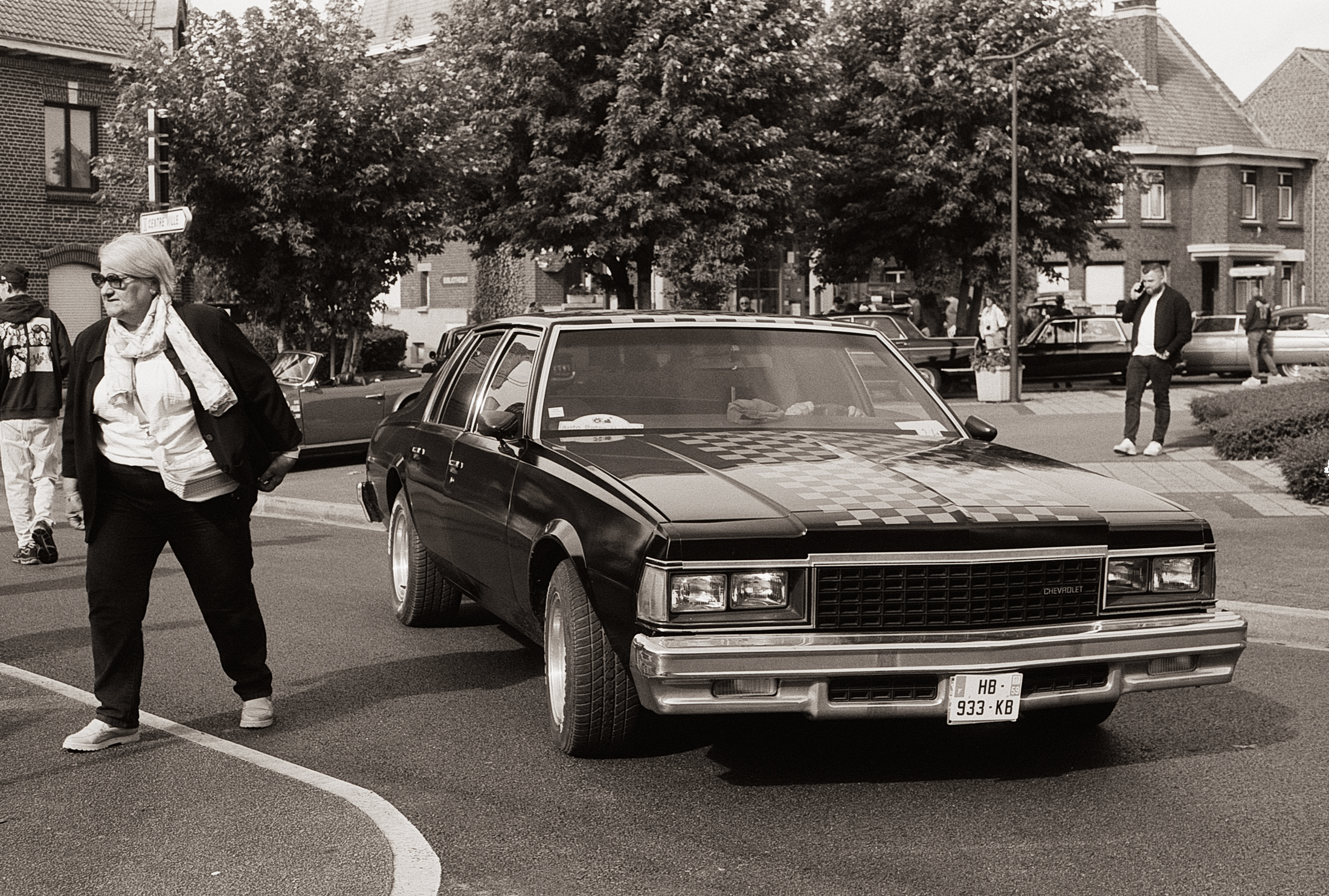 Une femme marche près d'une voiture noire avec un capot à damier, sur une route bordée d'arbres et de bâtiments résidentiels.