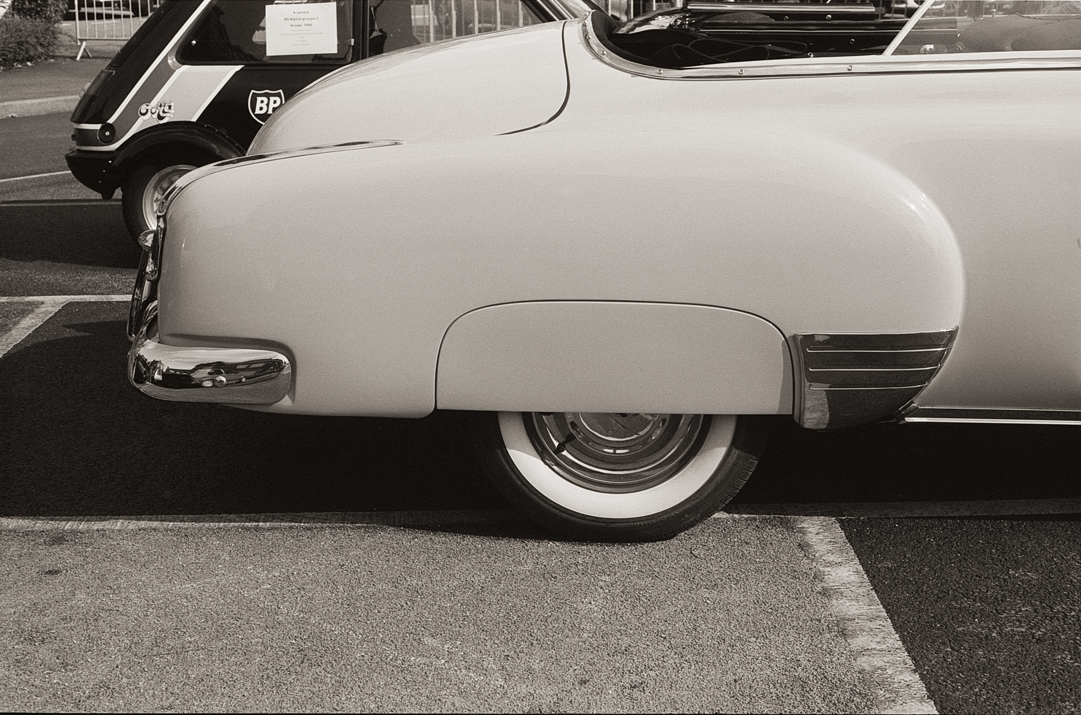 Vue latérale d'une voiture vintage blanche avec un design arrondi, montrant sa roue et un jante métallique brillant. En arrière-plan, une petite voiture noire est visible.
