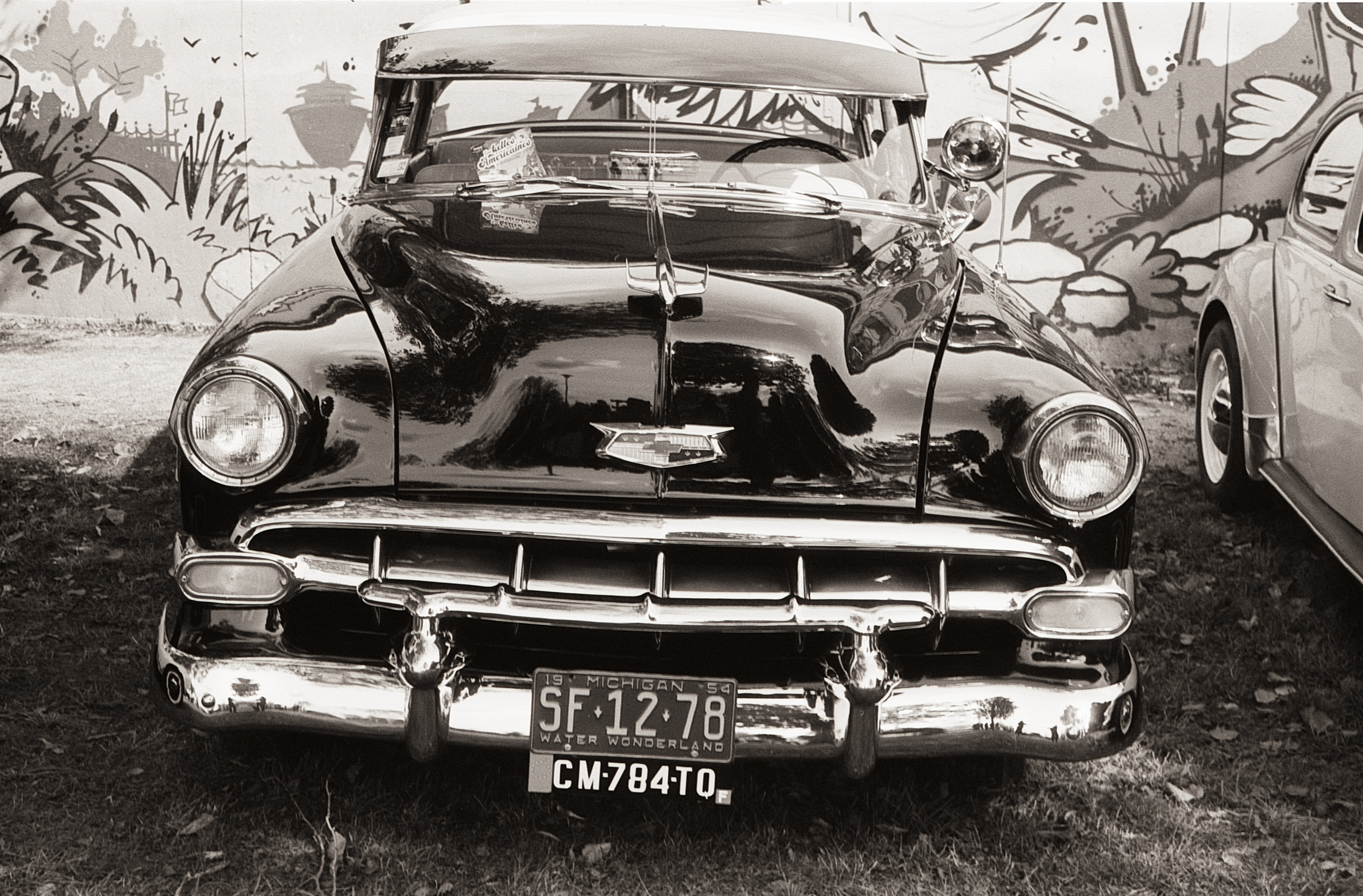 Vue frontal d'une voiture ancienne noire avec des chromes brillants, garée devant une fresque murale colorée.