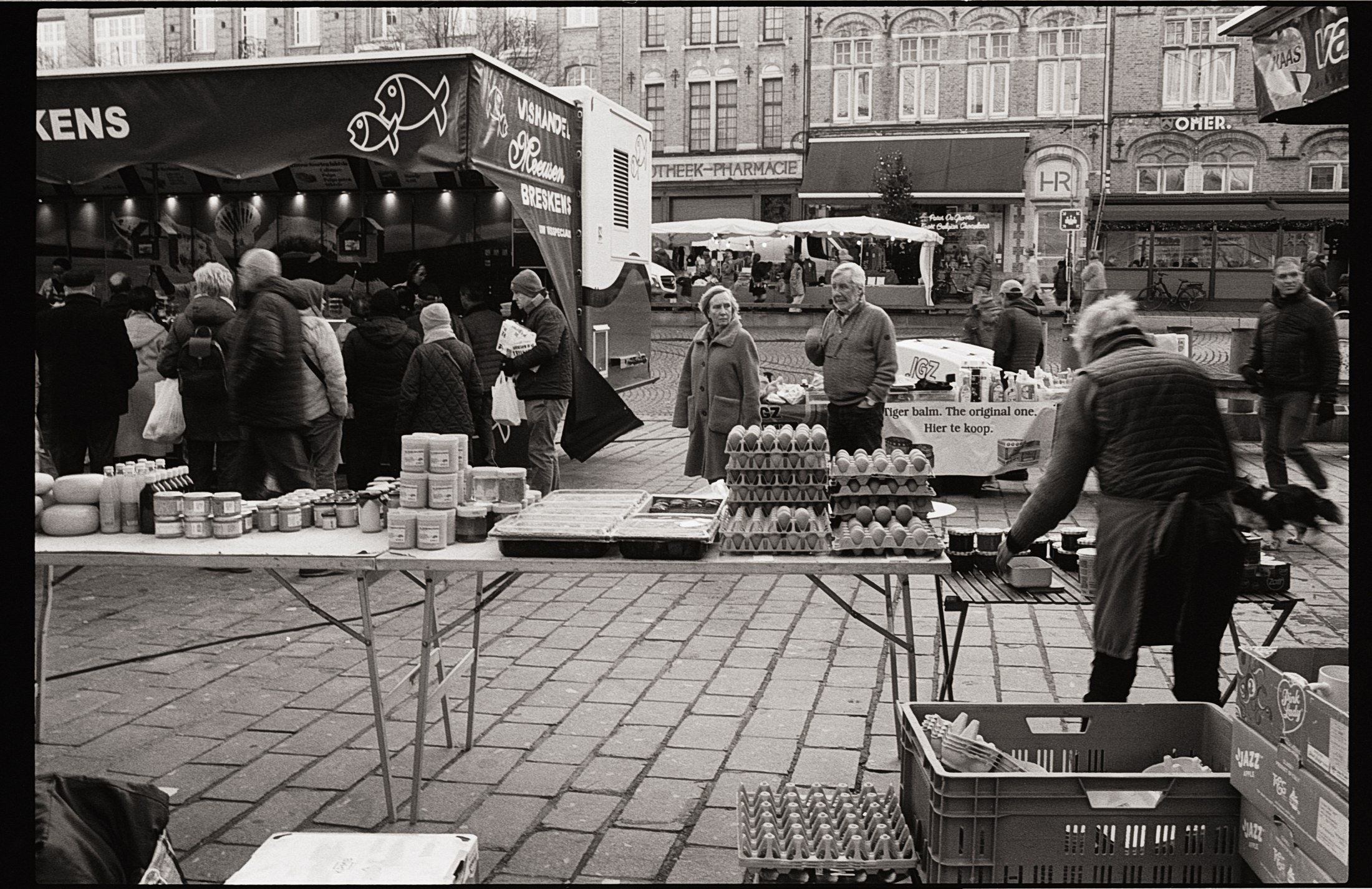 Une scène de marché en noir et blanc avec des gens faisant leurs courses, des étals de produits alimentaires comme des œufs et des conserves. Des personnes attendent près d'un stand de poissons visible en arrière-plan.