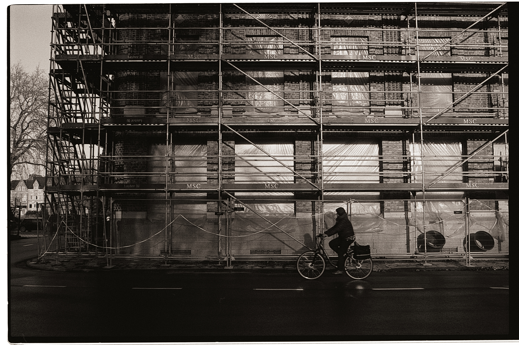 Un cycliste passe devant un bâtiment en construction, entouré d'échafaudages et de bâches. L'image est en noir et blanc, créant une atmosphère contemplative.