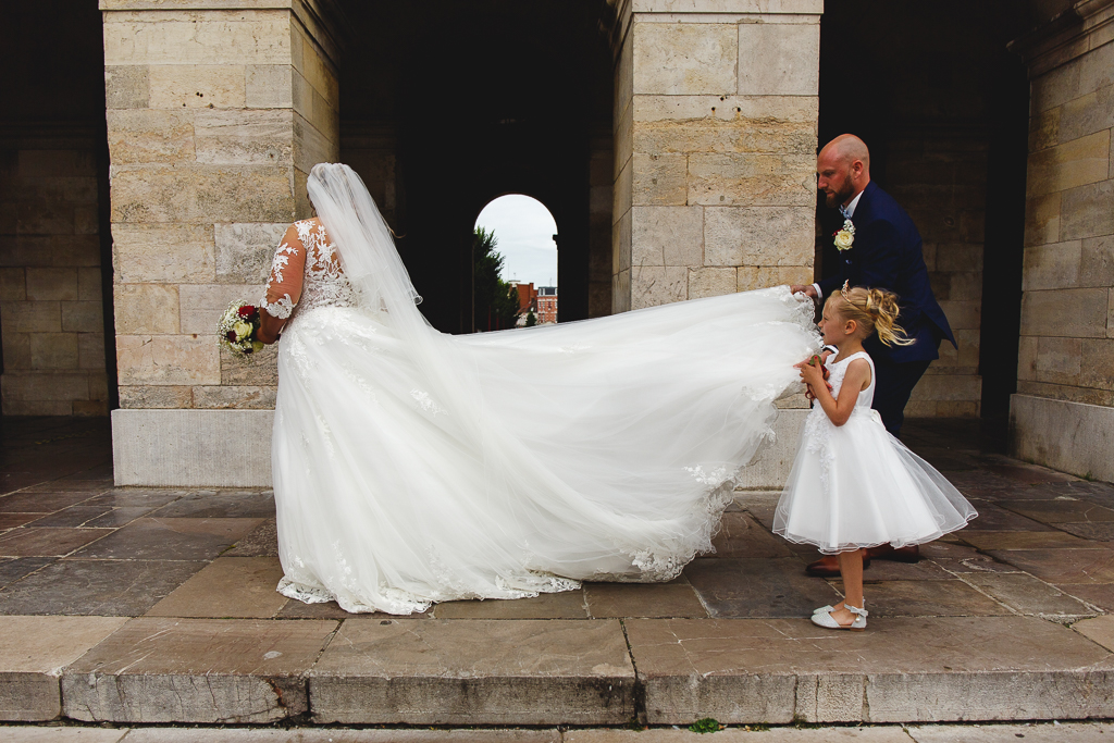 Une mariée en robe blanche avec un voile, tenant un bouquet, est suivie par un enfant de jeunes filles en robe blanche qui tient la traîne de sa robe. Un homme en costume bleu accompagne la mariée dans un cadre architectural.