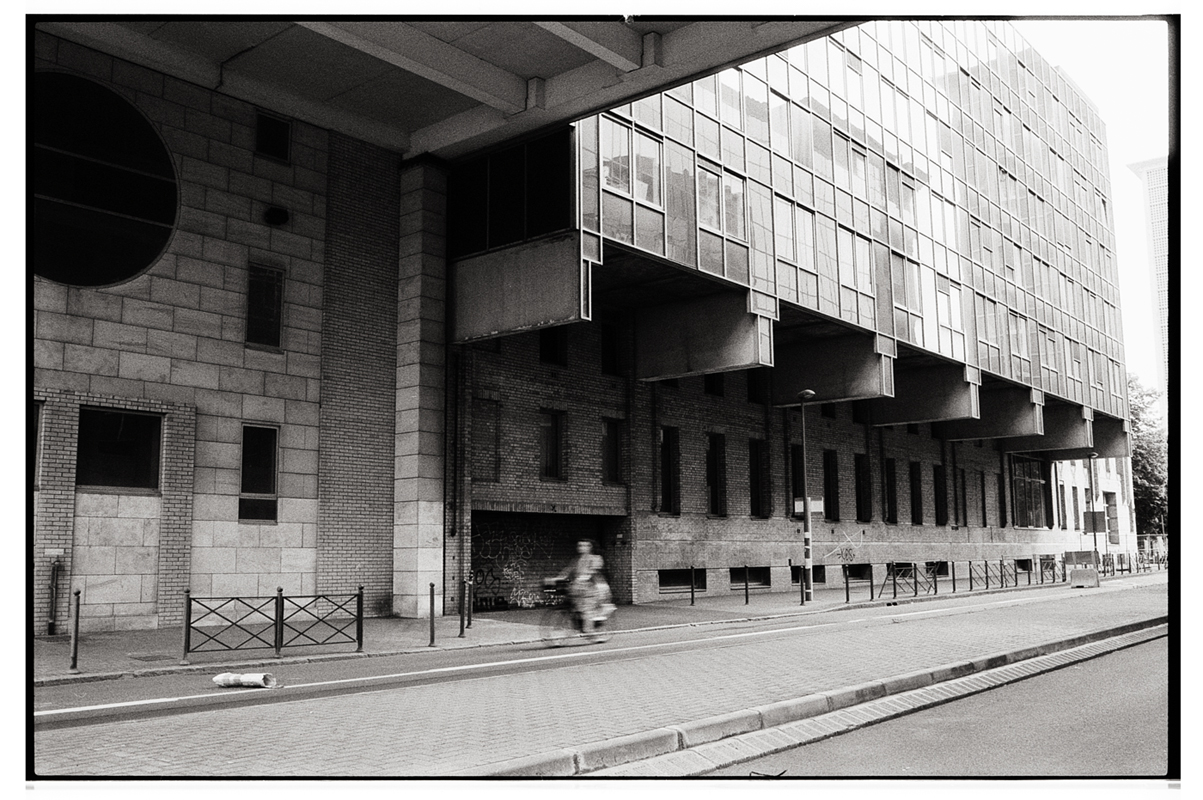 Une photographie en noir et blanc d'un bâtiment moderne avec des fenêtres en verre surélevées, situé au-dessus d'une structure en brique. Un cycliste passe en flou sur une rue presque déserte.